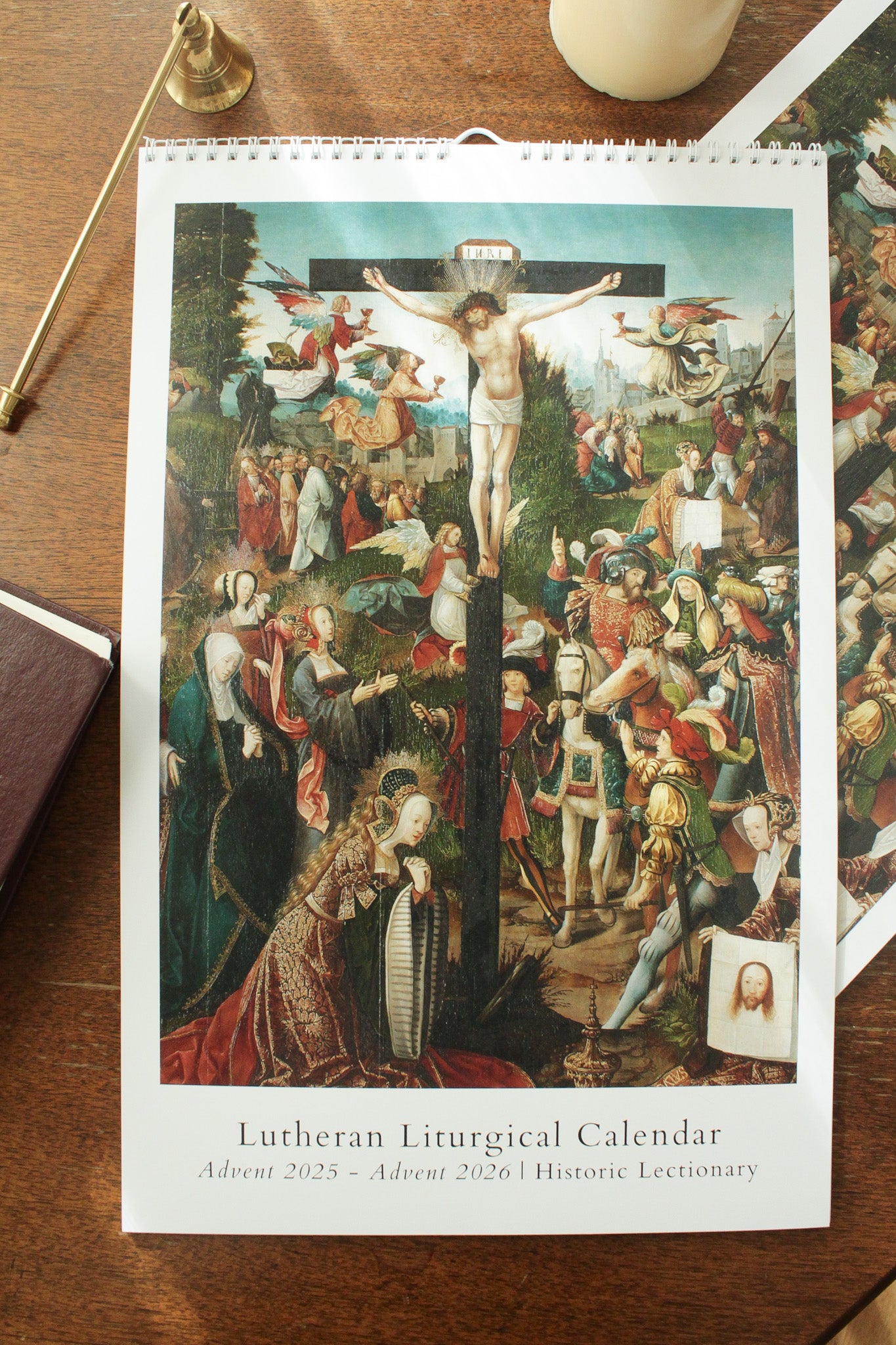 A Lutheran liturgical wall calendar for the years 2025-2026 featuring a colorful artwork of the crucifixion, with Sundays, feasts, commemorations, and lectionary readings marked on the pages.